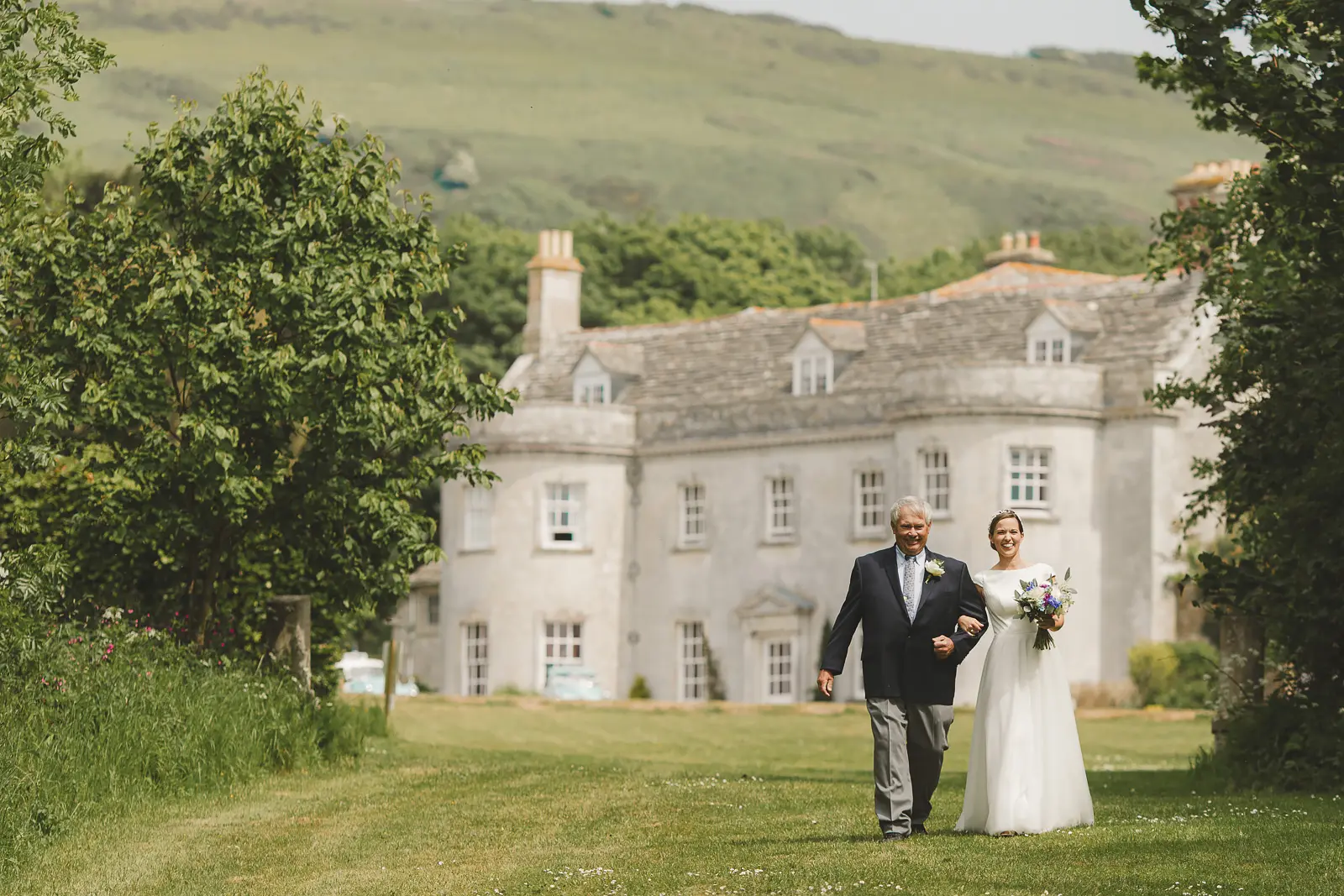 Bride walking with her father down the tree lined lane at Smedmore House towards the ceremony Dorset