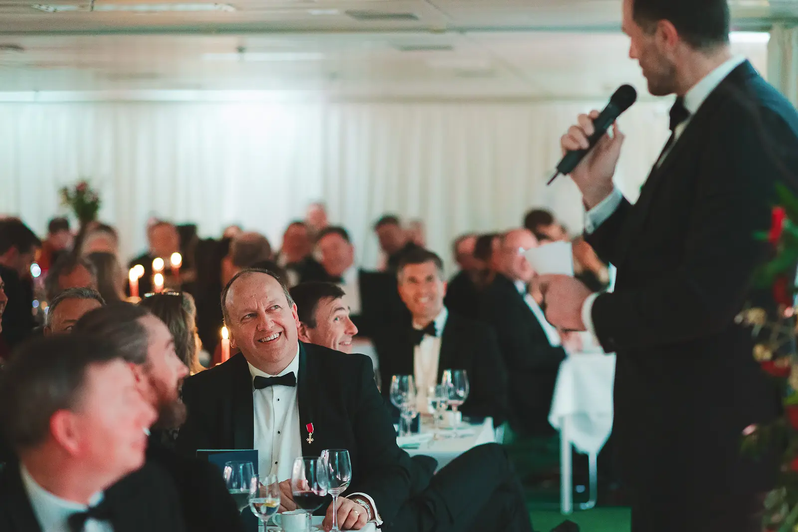 View from behind speaker with microphone addressing full formal dinner room, guests laughing at tables in candlelit setting