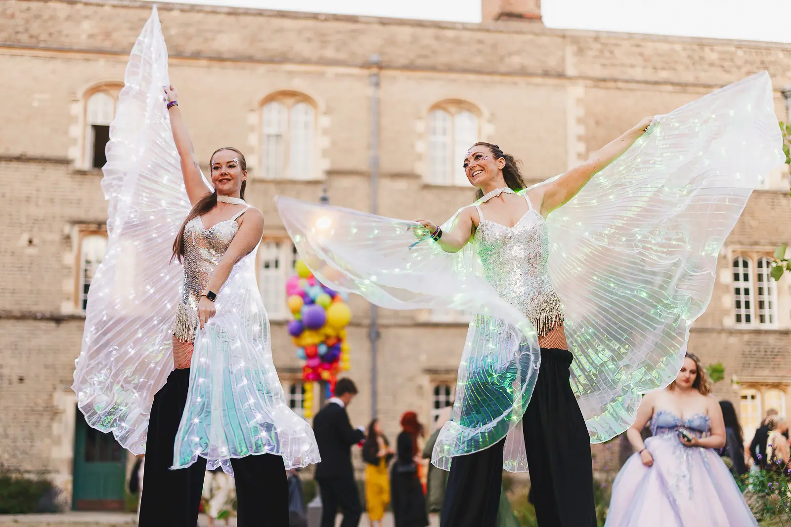 Two stilt performers with illuminated butterfly wings pose in front of the college building at a Cambridge summer ball