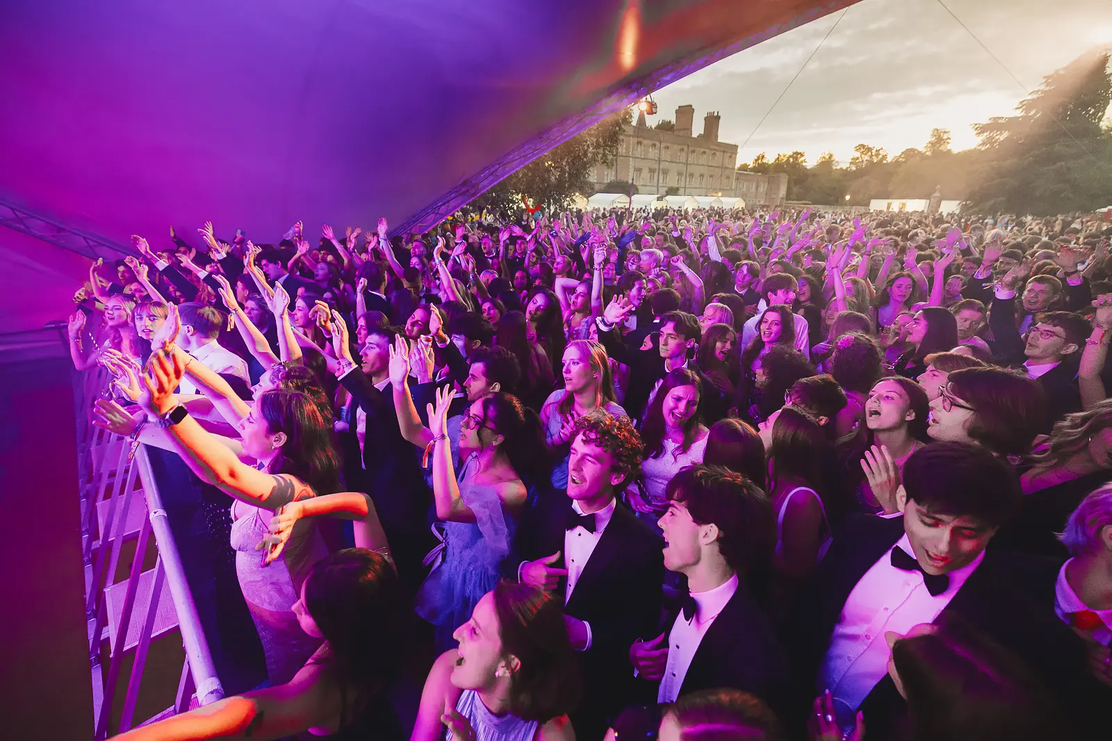 Thousands of formally dressed guests packed in front of the main stage at twilight with college buildings behind