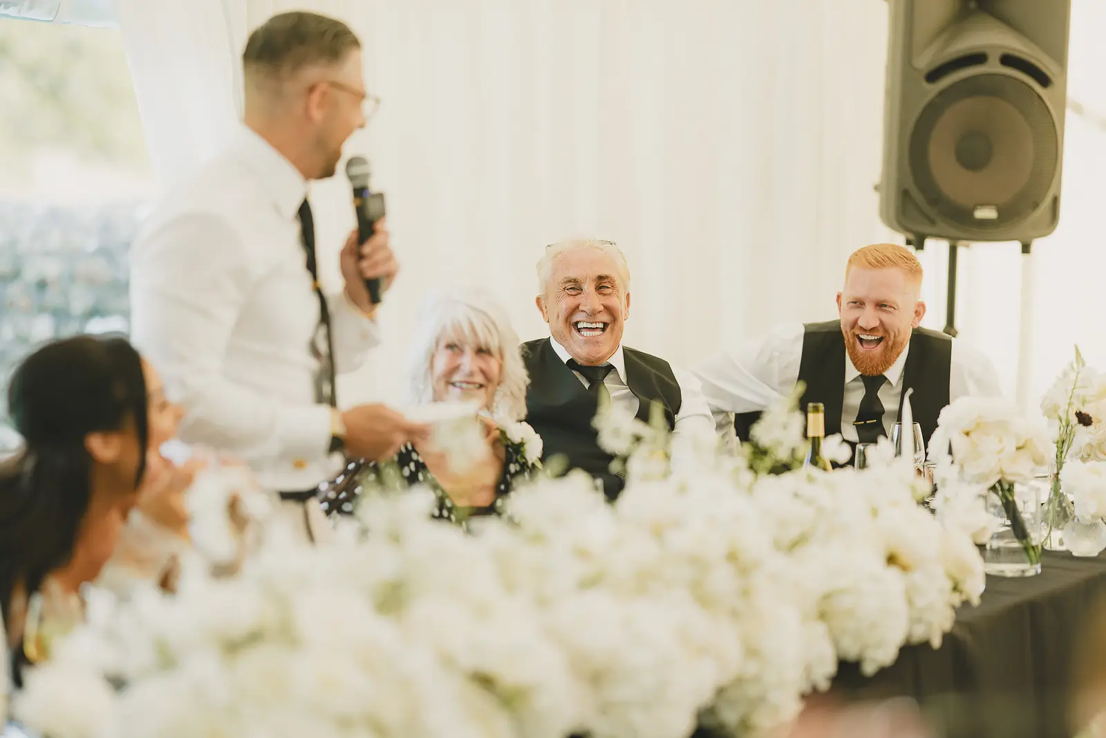 Top table guests laughing loudly during the wedding speeches in the marquee, white floral arrangements in the foreground