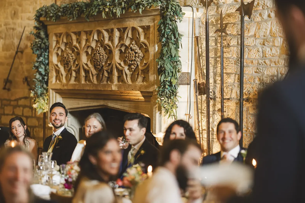 Guests and groom smiling at the top table with the ivy-draped Gothic carved stone fireplace behind during the wedding speeches in the tithe barn