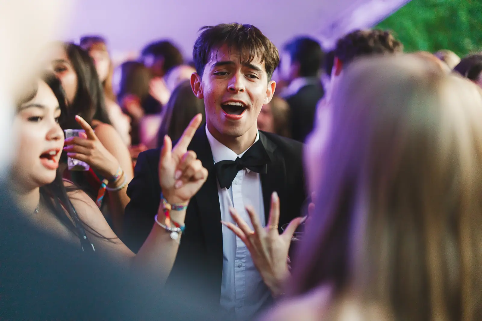 Guest in a tuxedo dancing and singing in a crowd under purple stage lights at a summer ball event