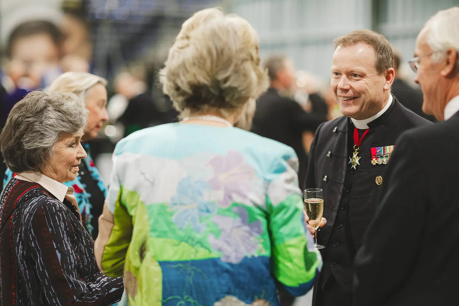 VIP guests in formal attire in conversation at evening reception, dignitary with medals holding champagne, documentary photography