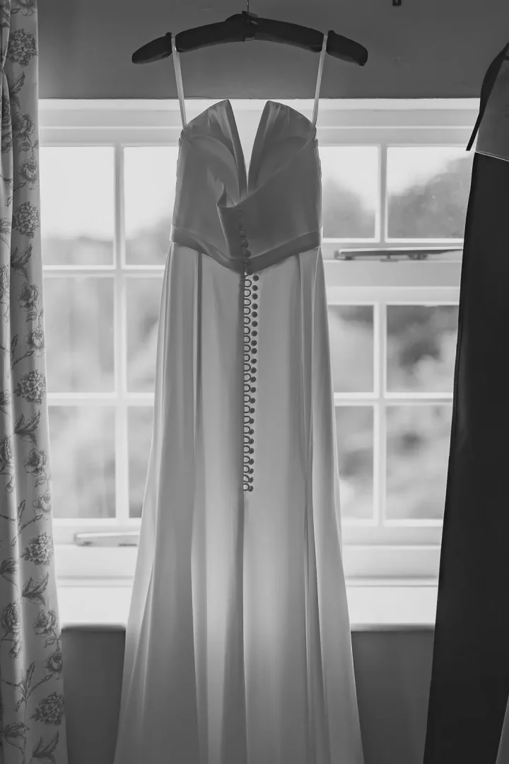 Bride's white wedding dress hanging in window light, button-back detail and cathedral length train visible