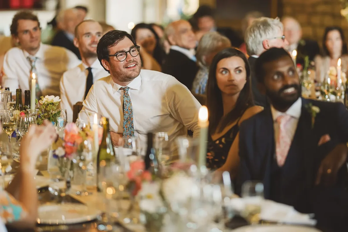 Wedding guests reacting with smiles at the reception tables during the speeches in the candlelit tithe barn at Hinton St Mary Estate