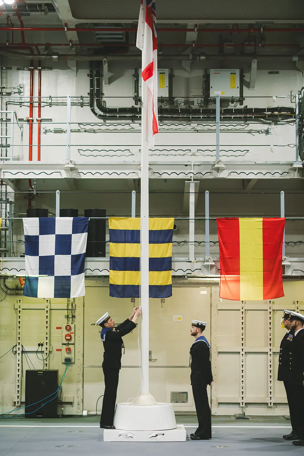 Sailors attending mast as White Ensign is raised inside hangar, naval signal flags displayed either side during formal ceremony