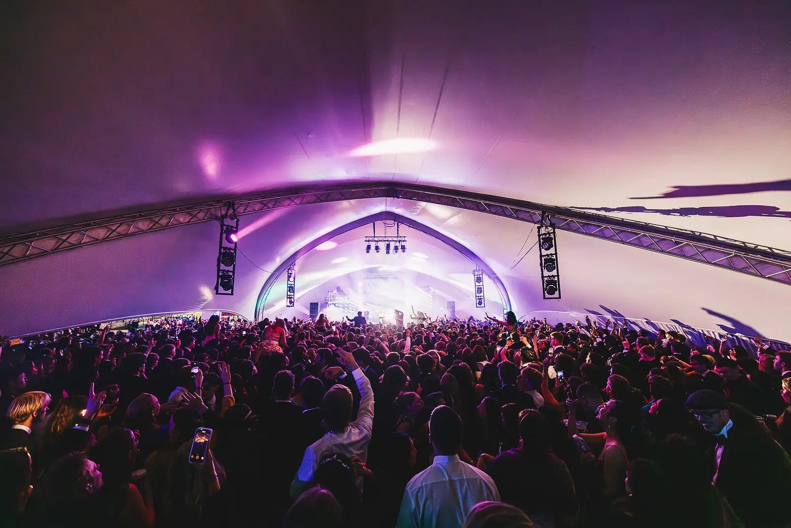Wide angle view of a dense crowd filling a large tent venue lit with purple stage lighting at a late-night summer ball