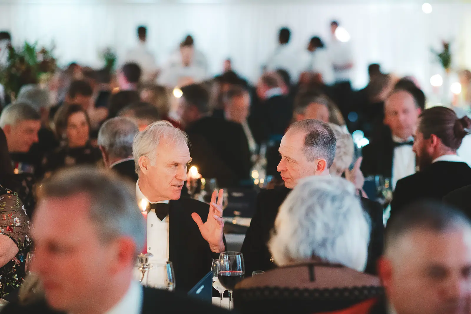 Wide reportage view of full formal dinner room, guests in conversation with naval stewards attending tables in background, candlelit atmosphere