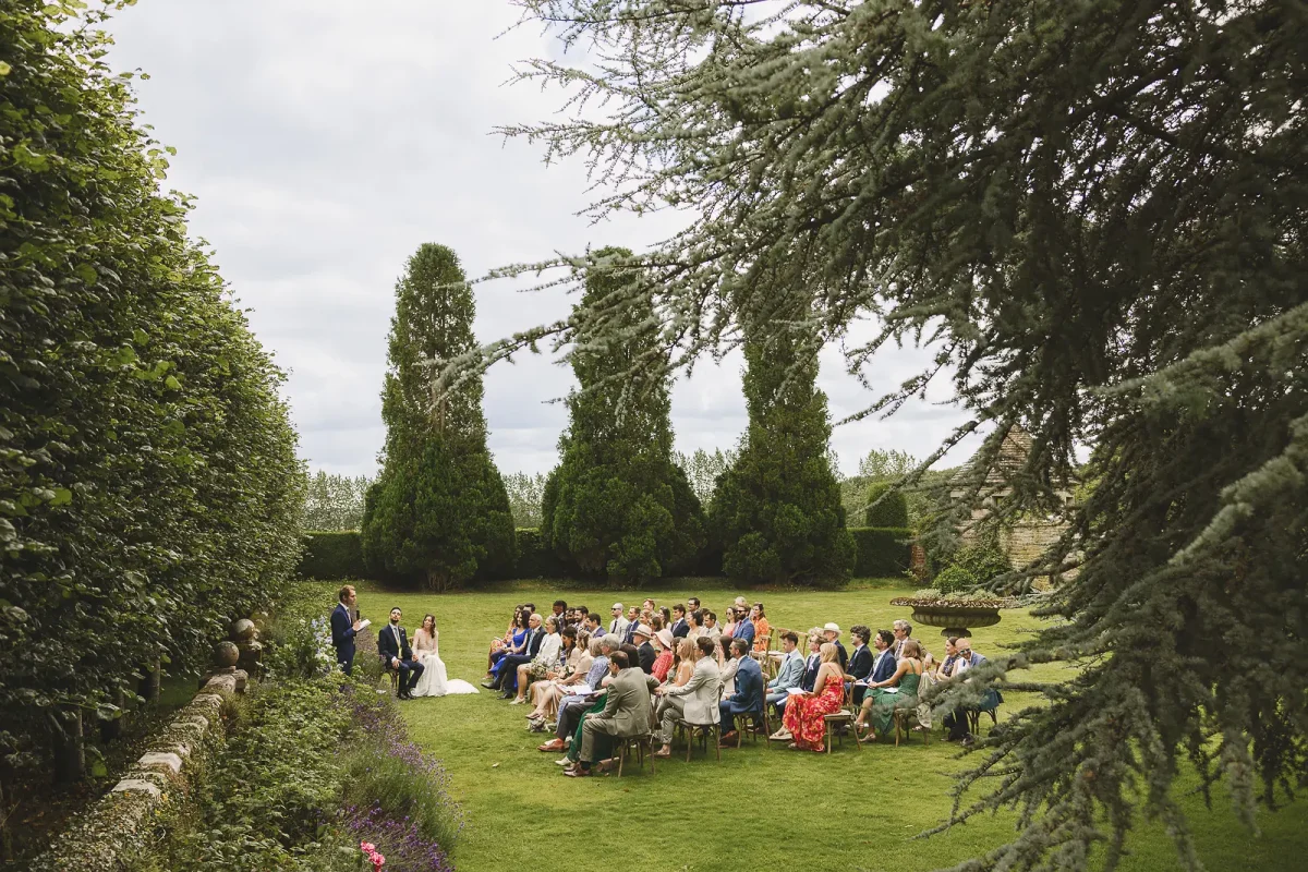 Wide view of the outdoor wedding ceremony with guests seated on the lawn surrounded by yew topiary at Hinton St Mary Estate