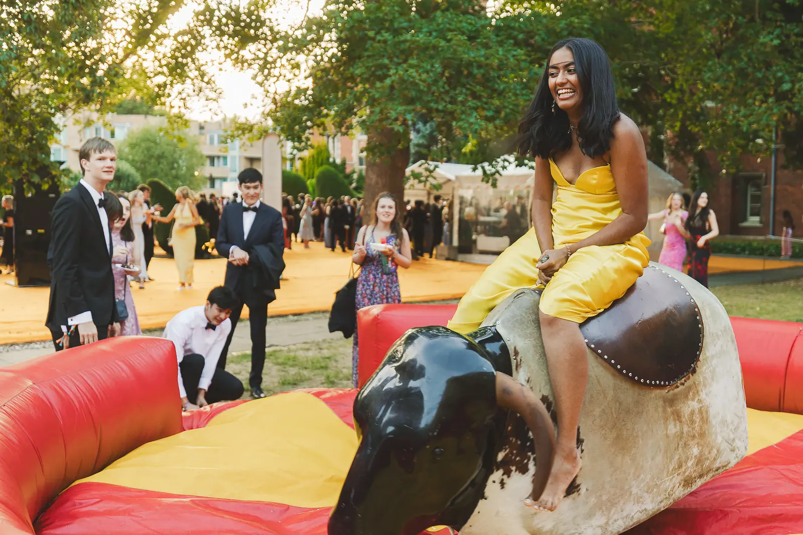 Guest in a yellow gown riding a mechanical bull at a summer ball with a crowd watching in the evening light