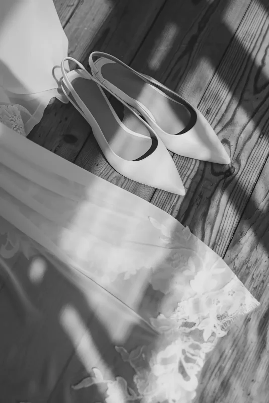 Black and white flat lay of pointed slingback bridal heels resting on lace wedding dress hem on wooden floor