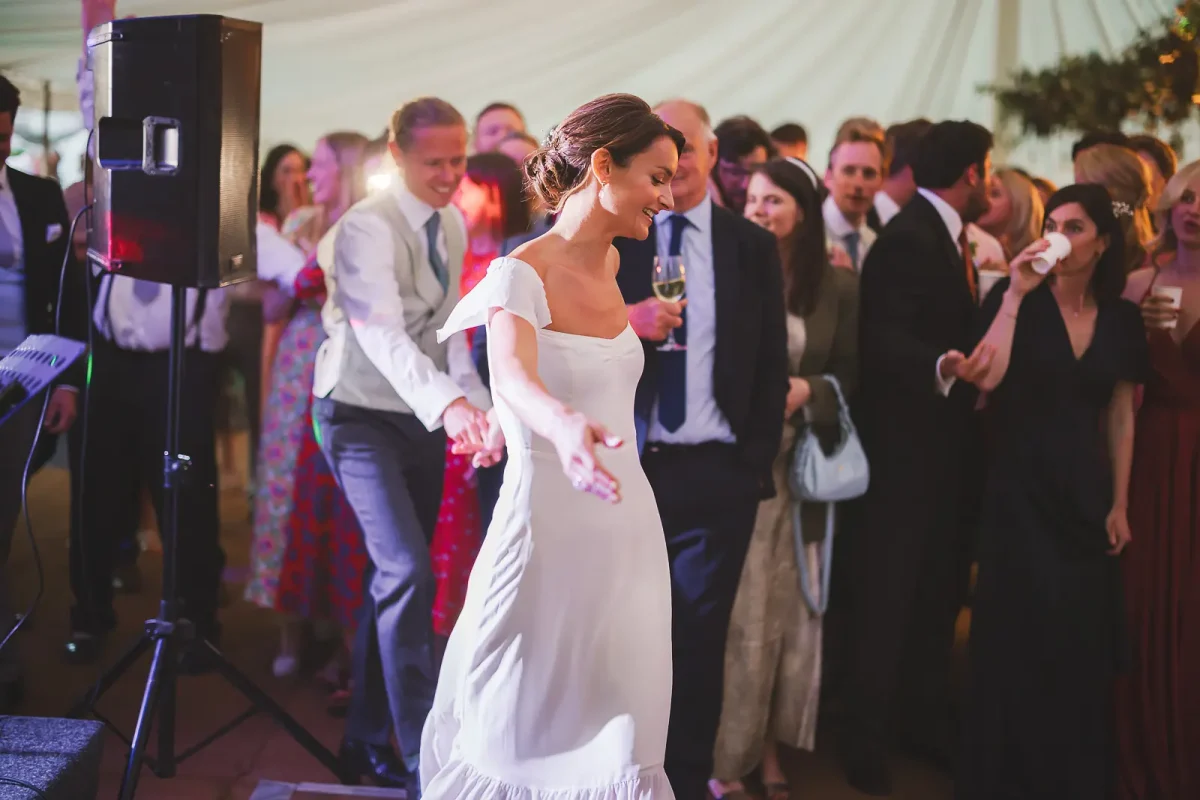 Bride in off-shoulder white dress dancing at the front of a packed dance floor at the marquee evening reception