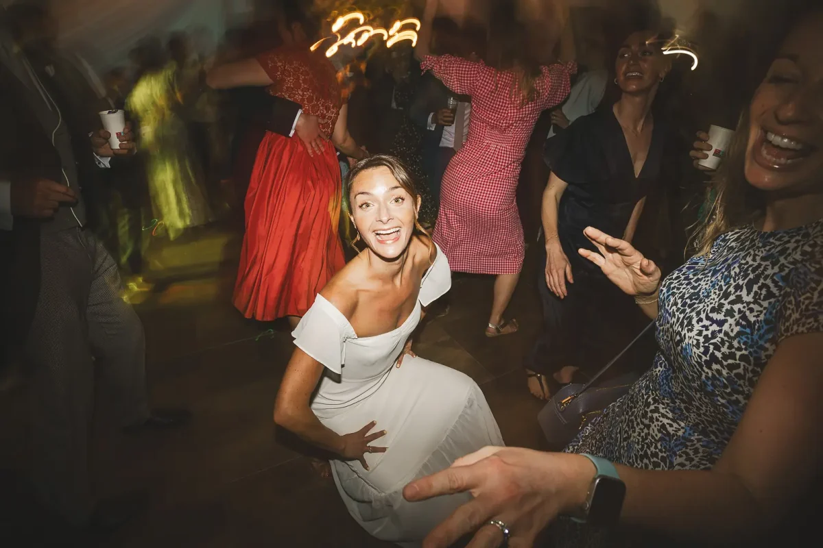 Bride in off-shoulder white dress grinning at the camera while dancing at the marquee evening party
