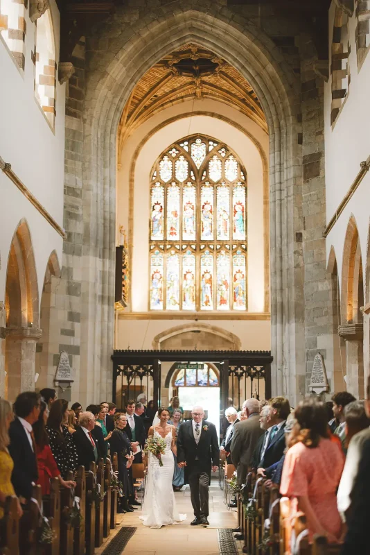 Bride and father walking down the aisle at Wimborne Minster church with stunning stained glass window behind them