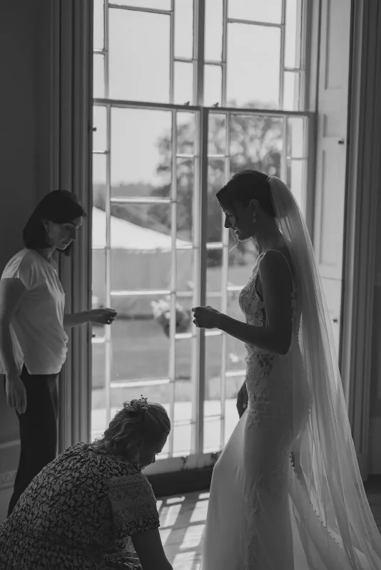 Black and white silhouette of bride in lace gown and veil being helped with final preparations at tall Georgian windows