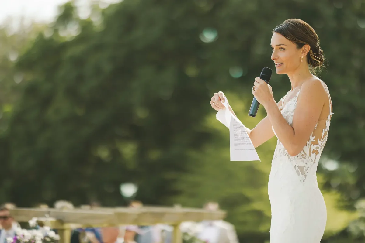 Bride in lace gown giving a speech into a microphone outdoors at the drinks reception with trees behind her
