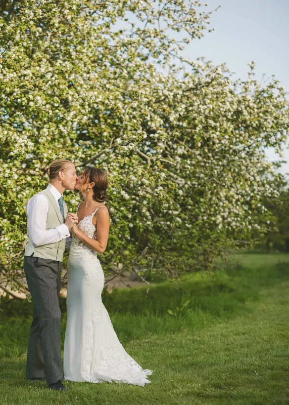 Bride and groom sharing a kiss beneath a full blossom tree in golden evening light at their summer wedding