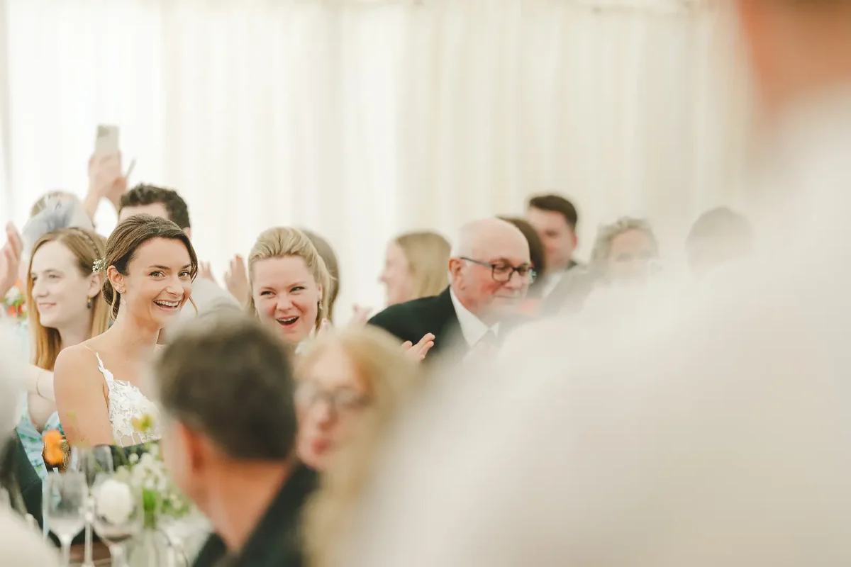 Bride in lace gown laughing with guests during wedding reception speeches inside a bright marquee