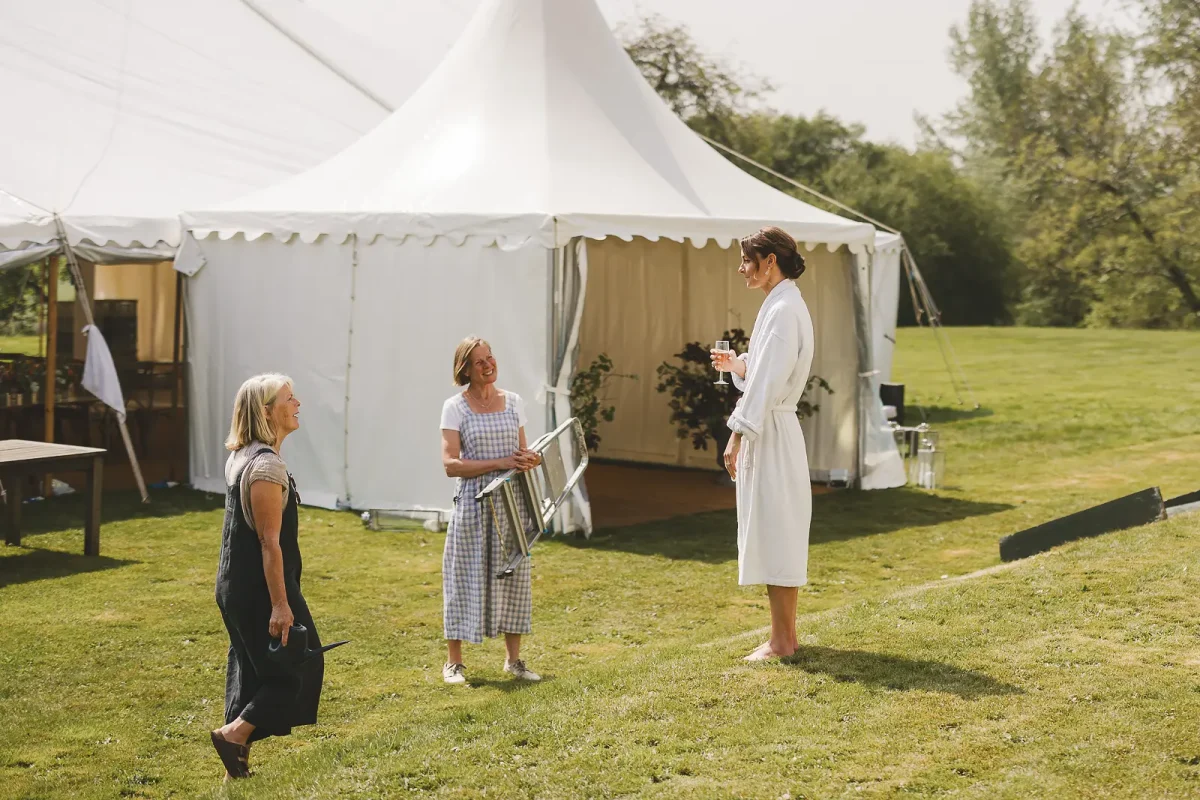 Bride in white robe with champagne chatting outside marquee during final setup on summer wedding morning