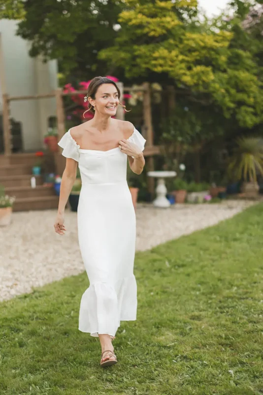 Bride in a white off-the-shoulder midi dress walking through the garden at the evening reception