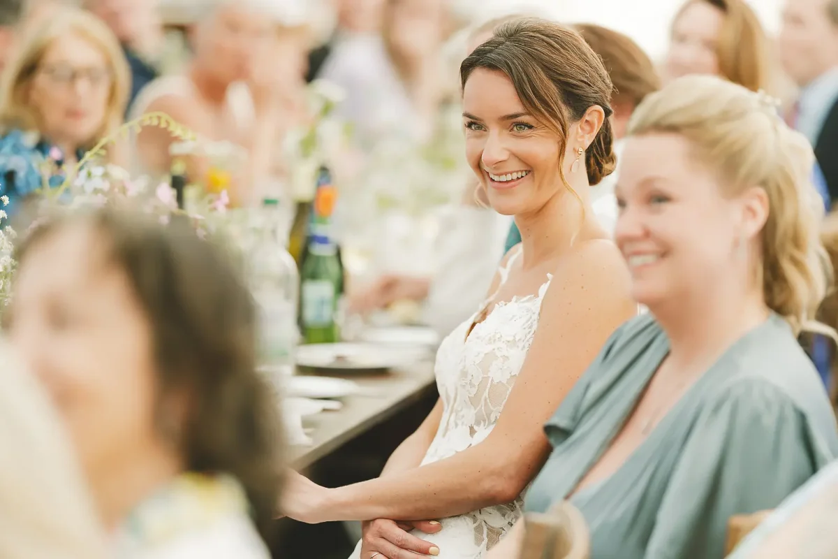 Bride in lace gown smiling warmly during wedding speeches at a summer marquee reception