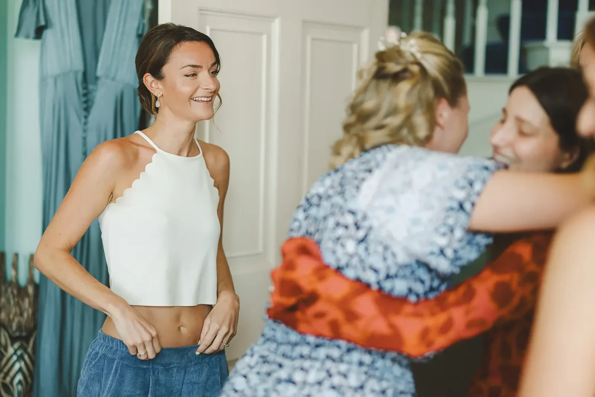 Bride smiling in white halter top while friends and bridesmaids gather around her during morning preparations