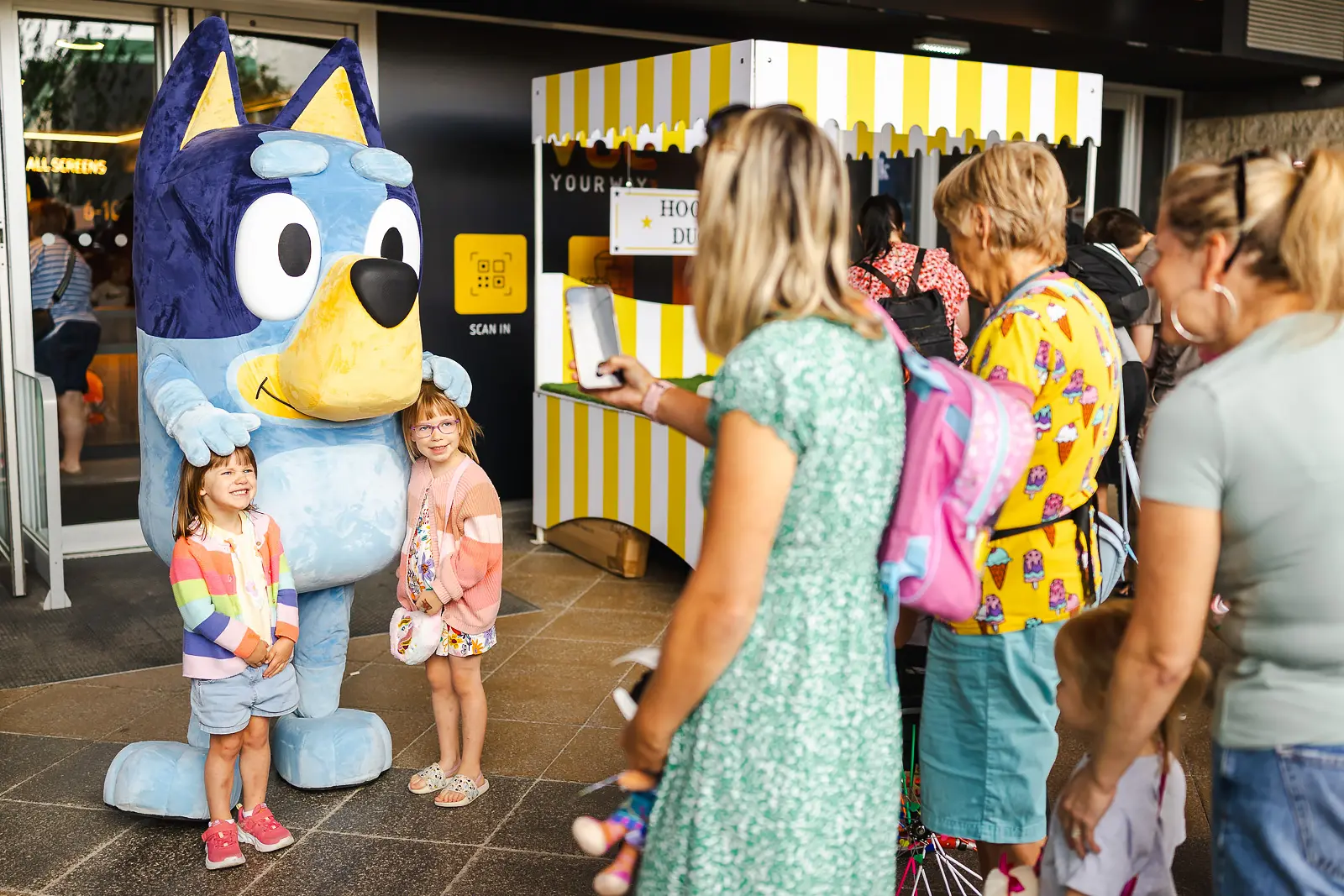 Two young girls posing with a costumed character at a public event in Poole, with families queuing in the background