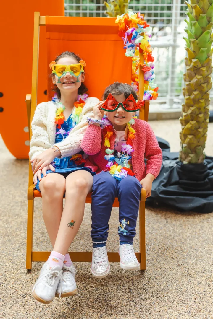 Two children sitting in an orange deckchair wearing novelty sunglasses and flower leis at a beach-themed event in Poole