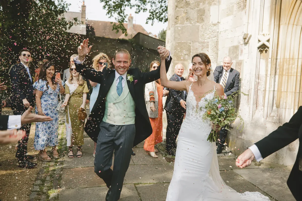 Newlyweds arms raised walking through a confetti cloud outside Wimborne Minster surrounded by colourfully dressed guests