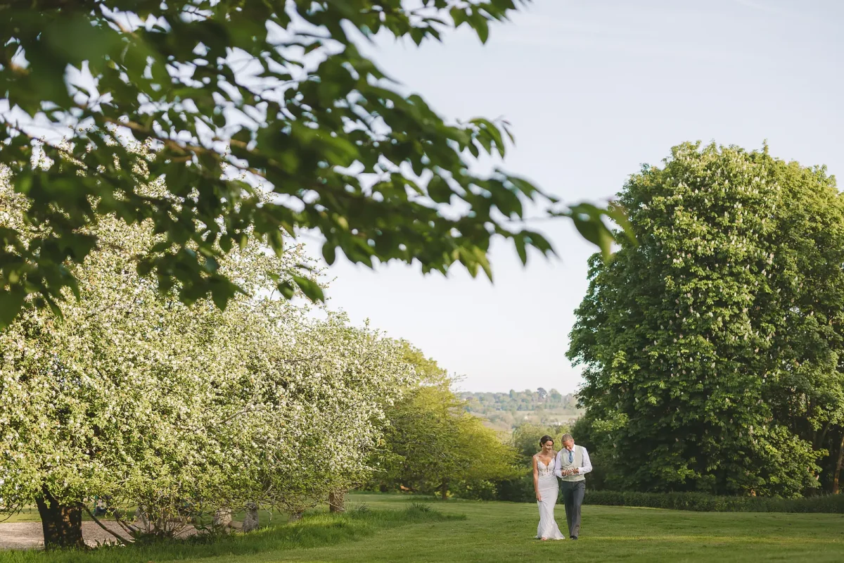 Bride and groom strolling beside flowering blossom trees in a green Dorset garden with countryside views beyond