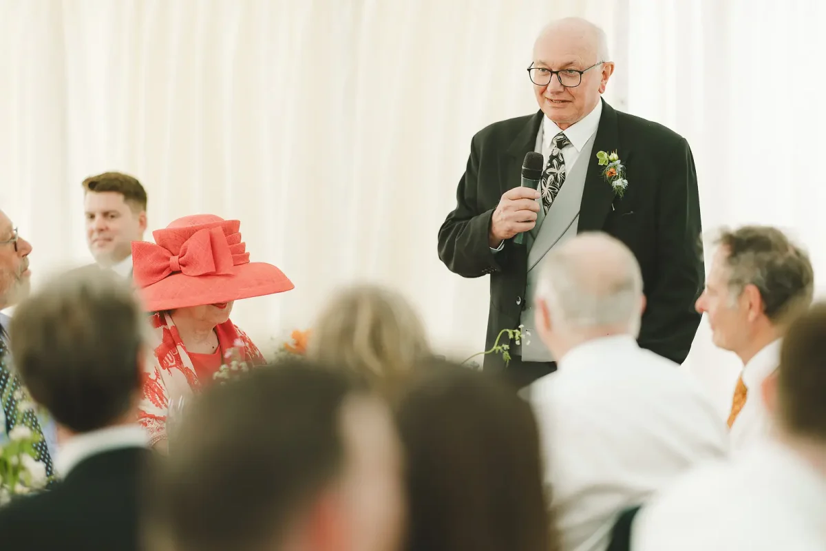 Father of the bride giving his speech inside the marquee with guests listening and the mother of bride beside him in a coral hat