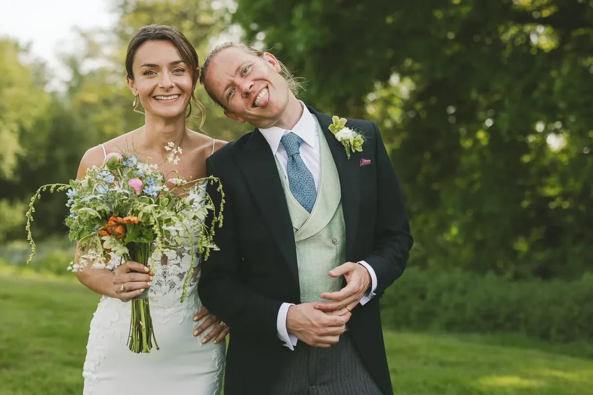 Bride smiling at camera while groom pulls a silly face in a lush green garden on their Dorset wedding day