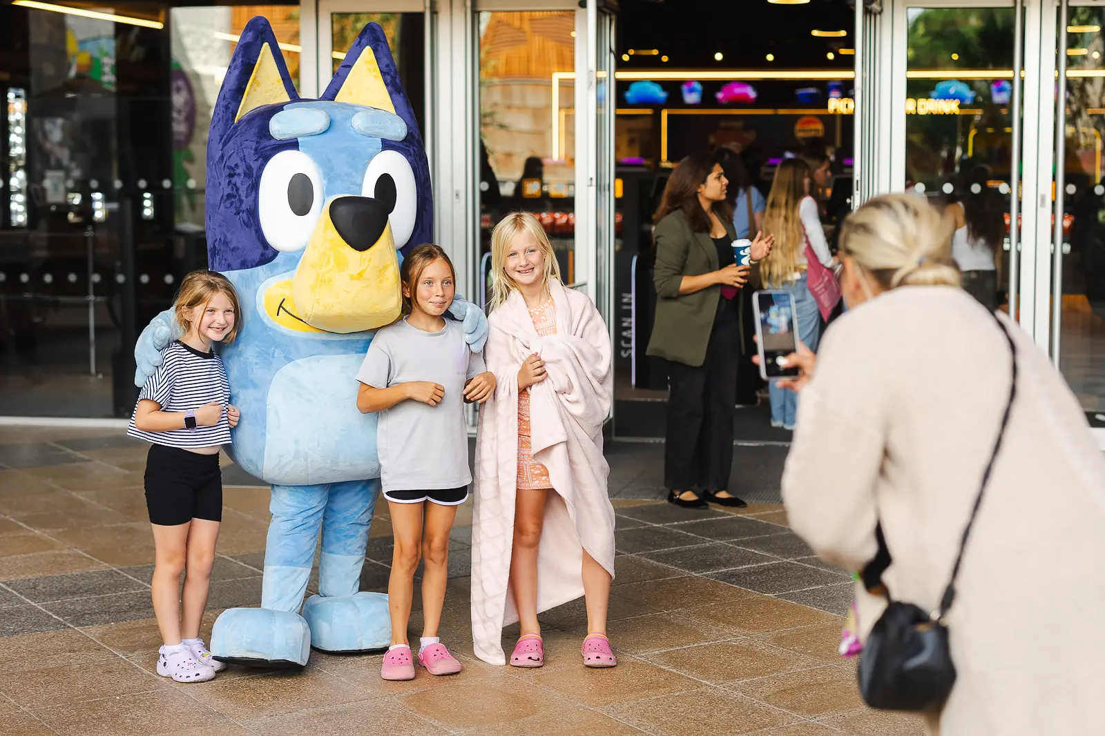 Three girls smiling as they pose with a costumed mascot outside a venue at a summer event in Poole, one wearing a beach towel