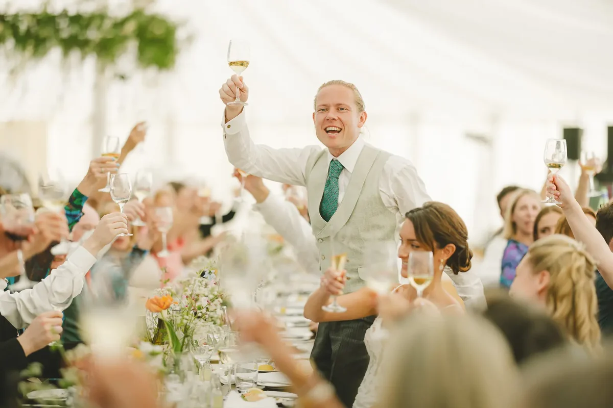 Groom raises his champagne glass to lead a toast as guests join in at the marquee wedding reception