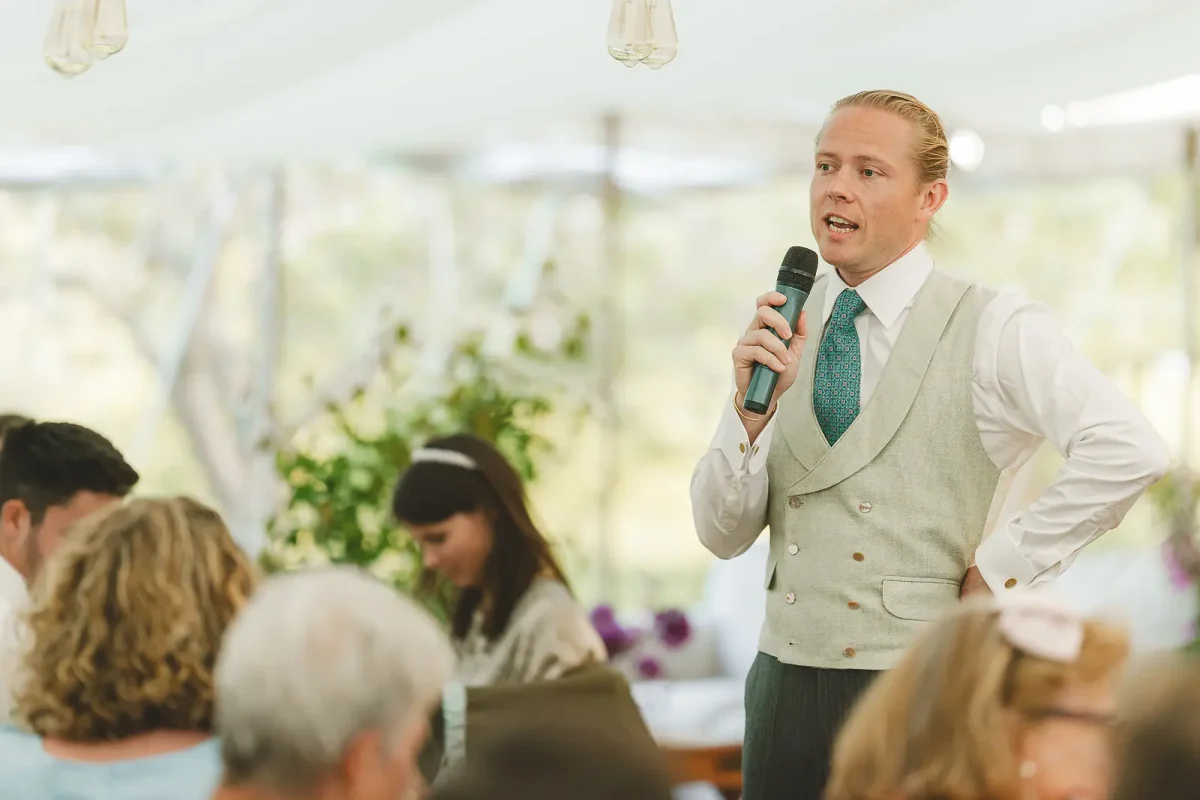 Groom addresses wedding guests with microphone in hand, standing at the reception inside a marquee