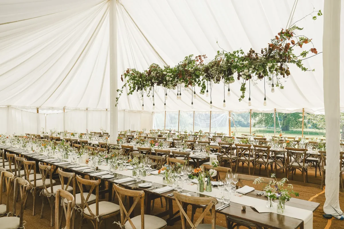 Marquee wedding reception interior with hanging greenery floral installation and long trestle tables set for dinner