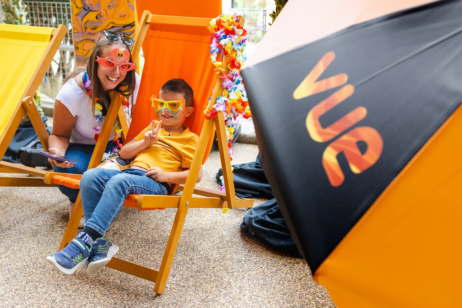 Mother crouching beside her son sitting in an orange deckchair with novelty sunglasses at a beach-themed event in Poole
