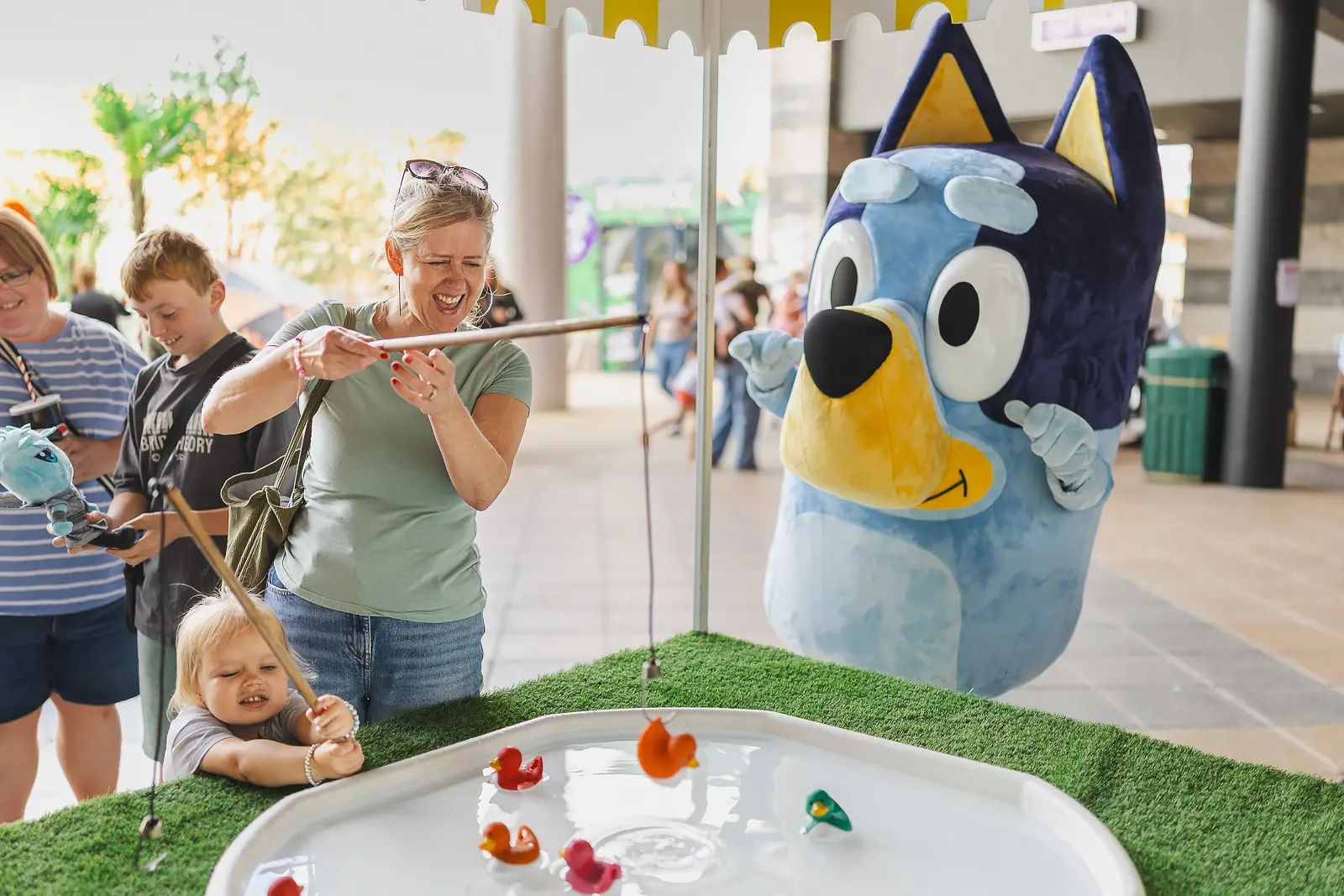 Woman and toddler concentrating on a duck pond fairground game beside a costumed character at an outdoor event in Poole