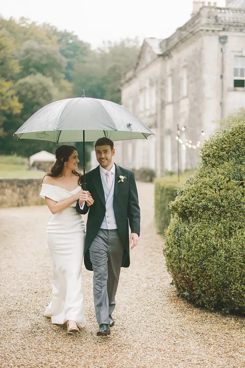 Bride and groom smiling under an umbrella during rainy wedding day portraits in Dorset
