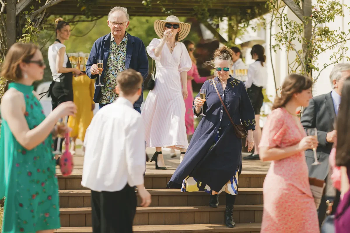 Colourfully dressed wedding guests with champagne mingling in a sunny garden during the outdoor drinks reception