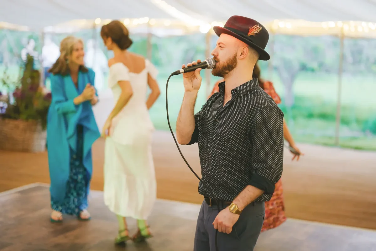 Wedding band singer performing with microphone as bride and guests dance behind him in the lit marquee