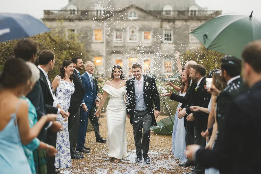 Bride and groom walking through confetti in the rain with guests and umbrellas in Dorset