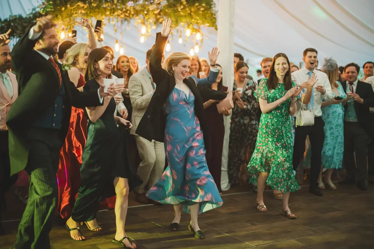 Wedding guests dancing energetically under floral chandelier and fairy lights at the evening marquee party