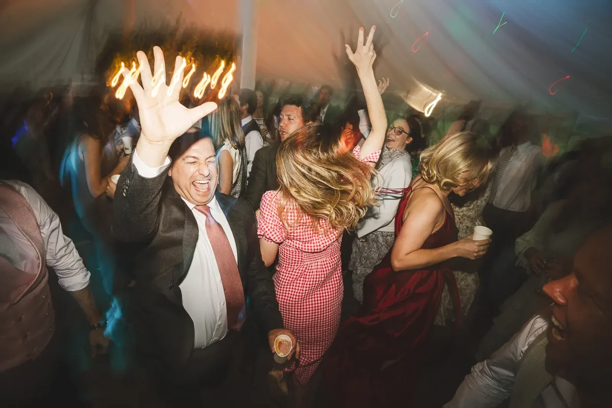 Wedding guests dancing energetically and throwing arms up at the marquee evening party