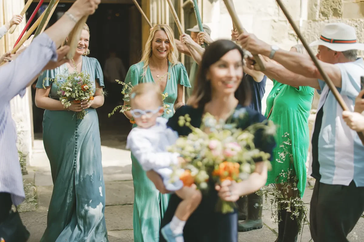 Bridesmaids and guests smiling as they file out of Wimborne Minster church with bouquets and a baby in the foreground