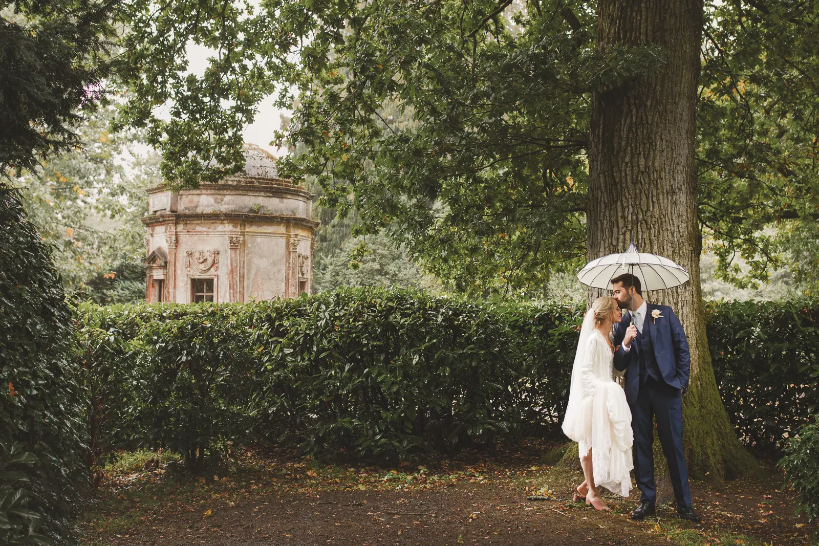 Bride and groom standing under an umbrella in the rain during a wet weather wedding portrait in a Dorset garden