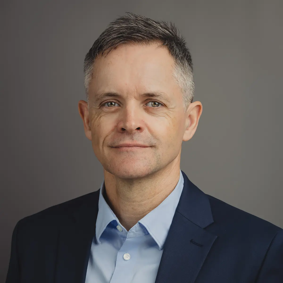 Professional headshot of a businessman in a navy jacket and blue shirt against a neutral studio-style background