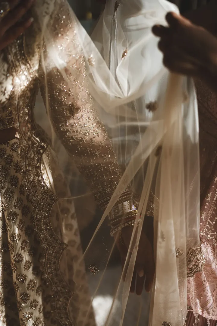 Atmospheric close-up detail of an intricately embroidered South Asian bridal lehenga with gold beadwork and bangles as a tulle veil is draped over the bride, Dorset wedding