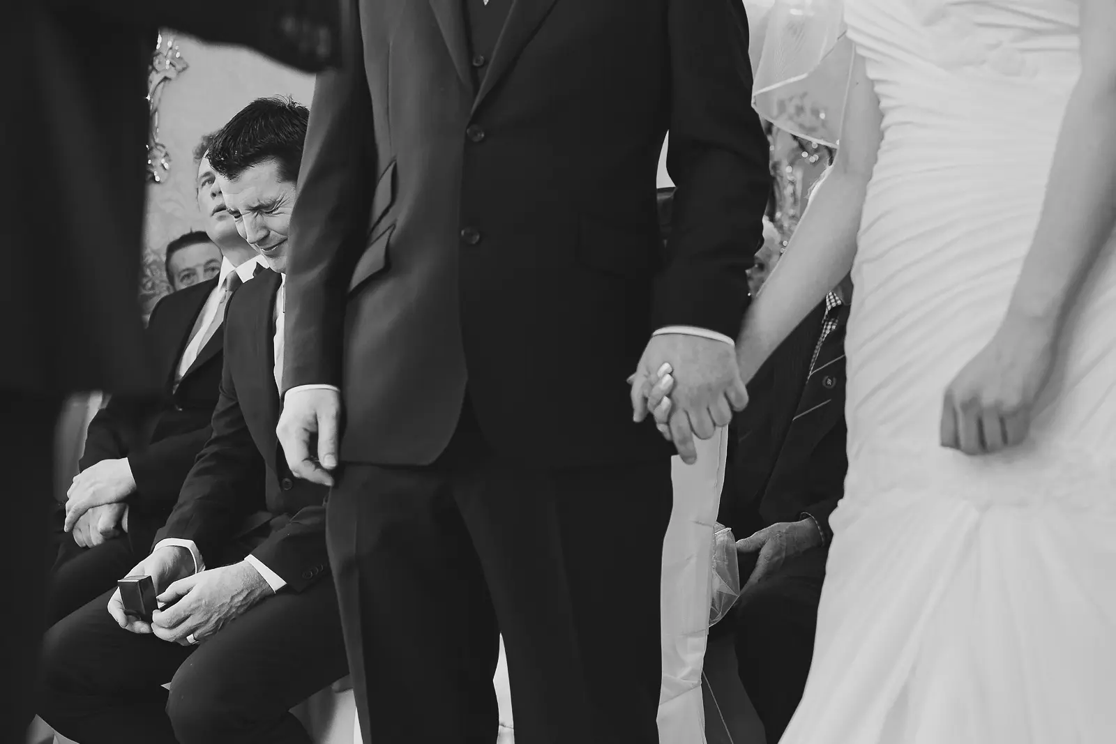 Candid black and white moment of best man laughing over ring box as bride and groom hold hands at wedding ceremony